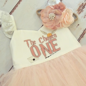 May include: A light pink tulle dress with the text "The Chosen One" in a Harry Potter theme. The dress has a cream-colored top with a decorative ruffle on the shoulder and a matching floral headband.