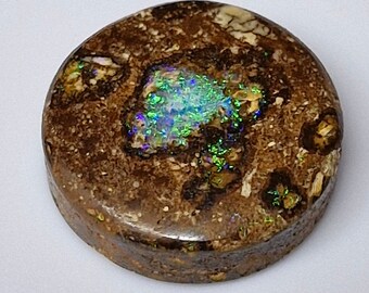 May include: A round, polished gemstone with a brown matrix and a central area of vibrant, iridescent opal. The opal displays flashes of green, blue, and purple. The gemstone has a smooth, rounded surface and a natural, earthy appearance.