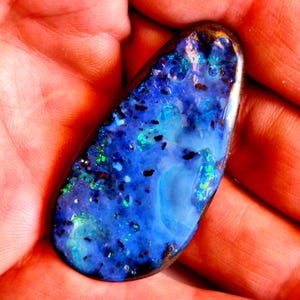 Puede incluir: Una piedra preciosa de ópalo pulida y alargada, con una vibrante exhibición de azules, verdes y toques de negro. La superficie de la piedra refleja la luz, mostrando un juego de colores. El ópalo se sostiene en una mano.