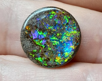 May include: A round, dark gray opal cabochon with a green, blue, and purple iridescent sheen. The opal has a rough, textured surface.
