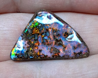 May include: A triangular opal gemstone displaying a spectrum of colours. The stone has blues, greens, oranges, and purples, set within a dark, textured matrix. The polished opal reveals an iridescent play-of-colour, a stunning natural gem.
