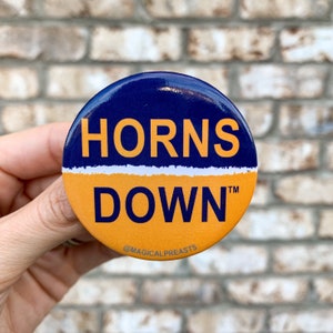 May include: A round button with a navy blue and orange design. The top half is navy blue with the word "HORNS" in orange. The bottom half is orange with the word "DOWN" in orange. The text "@MAGICALPREASTS" is at the bottom.