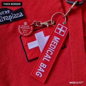 May include: A red fabric bag featuring a white embroidered cross and a rectangular tag with the text MEDICAL BAG. A round red tag attached by a metal clip reads Steroid Dependent CALL 911. A black patch with white text reads SURF Ilvaplana.