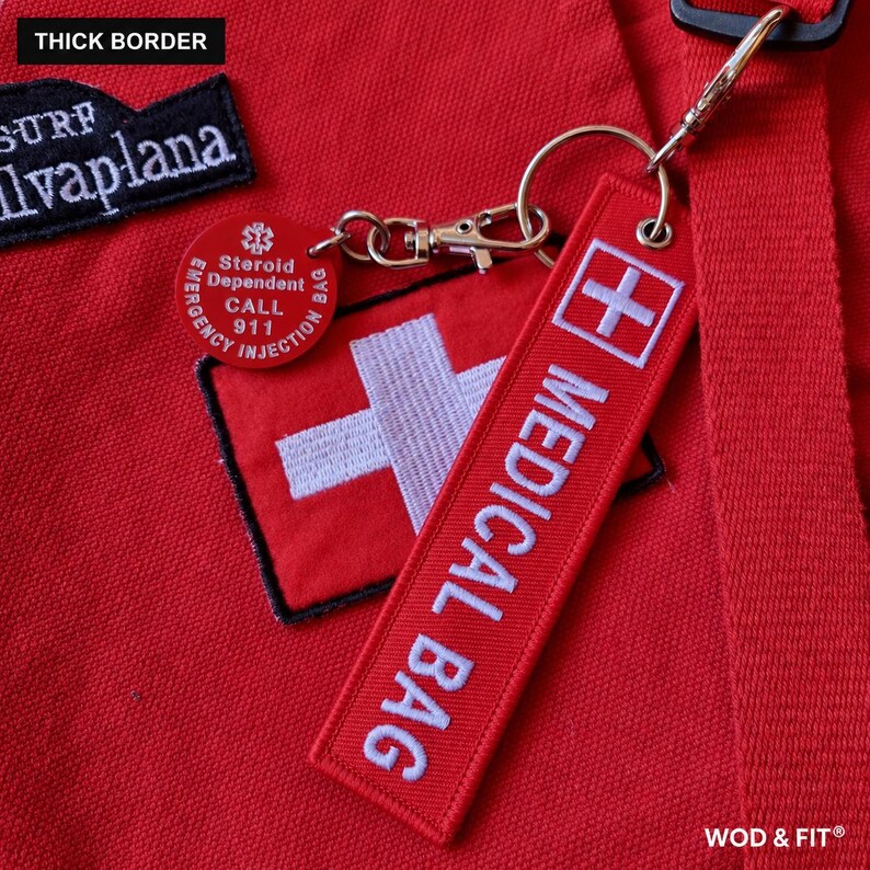 May include: A red fabric bag featuring a white embroidered cross and a rectangular tag with the text MEDICAL BAG. A round red tag attached by a metal clip reads Steroid Dependent CALL 911. A black patch with white text reads SURF Ilvaplana.