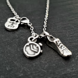 Puede incluir: Collar de plata con tres colgantes: una mancuerna con el texto "WOD & FIT", un cronómetro y una zapatilla de correr.