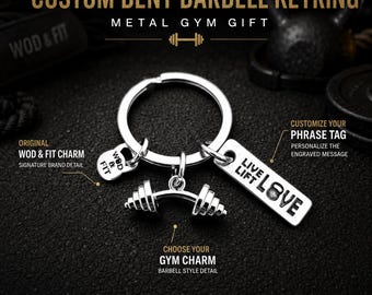 May include: A silver-toned metal keyring featuring a circular ring, a barbell-shaped charm, a small kettlebell charm engraved with WOD & FIT, and a rectangular tag engraved with the text LIVE LIFT LOVE, designed as a customizable gym accessory.