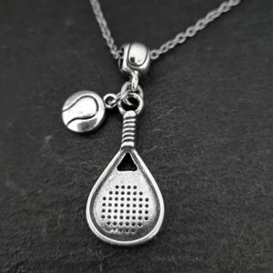 May include: Silver necklace with a charm of a paddle tennis racket and a tennis ball. The necklace is on a dark gray background.