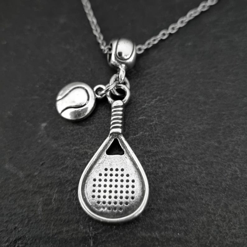 May include: Silver necklace with a charm of a paddle tennis racket and a tennis ball. The necklace is on a dark gray background.