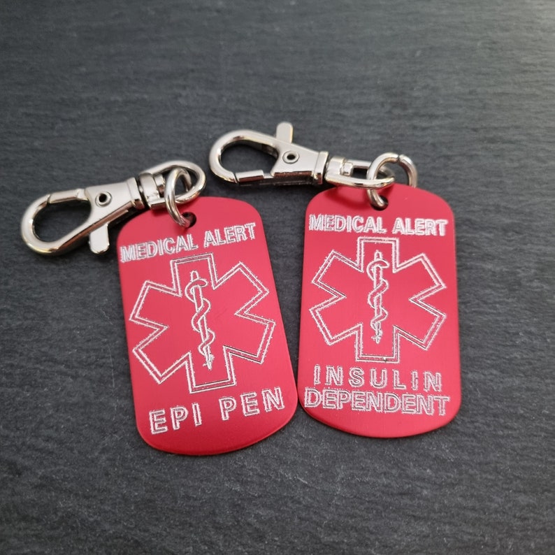 May include: Two red metal medical alert tags with silver keychains. The tag on the left says "Medical Alert" with a star of life symbol and "Epi Pen". The tag on the right says "Medical Alert" with a star of life symbol and "Insulin Dependent".
