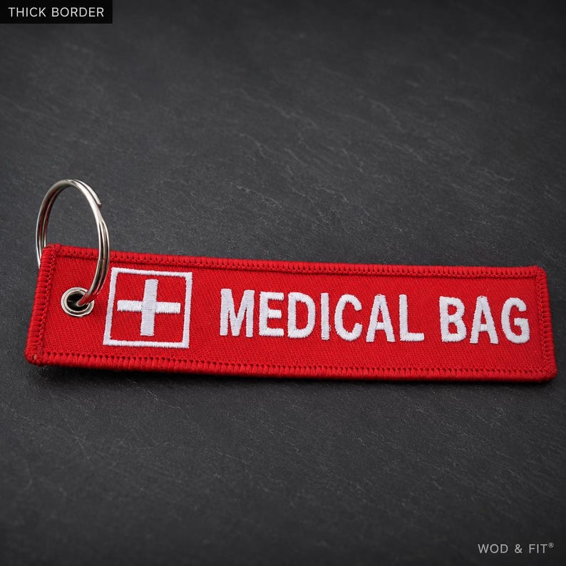 May include: A rectangular red fabric keychain with a white embroidered cross symbol and the text MEDICAL BAG in white capital letters. The keychain features a silver metal split ring attached through a reinforced eyelet on the left side.