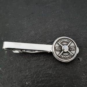Può includere: Un fermacravatta argentato con un design a peso per bilanciere. Il peso è rotondo e presenta la scritta "BARBELL" in alto e "STANDARD" in basso. Mostra "20,4 KGS" e "45 LBS". Un accessorio elegante per gli appassionati di fitness.