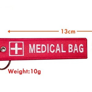 K&ouml;nnte beinhalten: Roter Stoffschl&uuml;sselanh&auml;nger mit einem wei&szlig;en Kreuz und dem Text "MEDICAL BAG" in wei&szlig;. Der Schl&uuml;sselanh&auml;nger ist 13 cm lang und 2,8 cm breit und wiegt 10 Gramm.