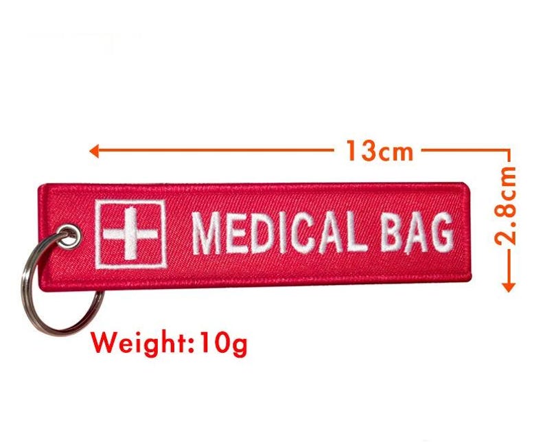 K&ouml;nnte beinhalten: Roter Stoffschl&uuml;sselanh&auml;nger mit einem wei&szlig;en Kreuz und dem Text "MEDICAL BAG" in wei&szlig;. Der Schl&uuml;sselanh&auml;nger ist 13 cm lang und 2,8 cm breit und wiegt 10 Gramm.