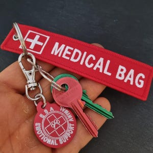 K&ouml;nnte beinhalten: Roter Schl&uuml;sselanh&auml;nger mit einem wei&szlig;en "MEDICAL BAG"-Etikett und einem wei&szlig;en Kreuz. An dem Schl&uuml;sselanh&auml;nger ist ein runder roter Anh&auml;nger mit einem wei&szlig;en Pfotenabdruck und dem Text "ESA Emotional Support Animal" befestigt.
