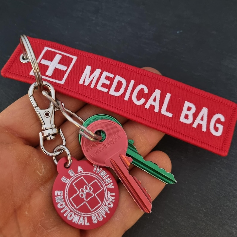 K&ouml;nnte beinhalten: Roter Schl&uuml;sselanh&auml;nger mit einem wei&szlig;en "MEDICAL BAG"-Etikett und einem wei&szlig;en Kreuz. An dem Schl&uuml;sselanh&auml;nger ist ein runder roter Anh&auml;nger mit einem wei&szlig;en Pfotenabdruck und dem Text "ESA Emotional Support Animal" befestigt.