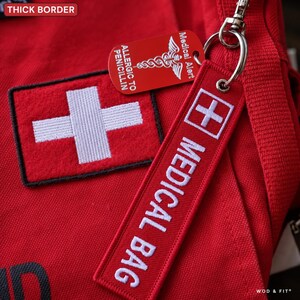May include: A red medical bag featuring a white cross patch and a red fabric keychain embroidered with white text reading MEDICAL BAG. A red metal medical alert tag with a white caduceus symbol and the text ALLERGIC TO PENICILLIN is attached.