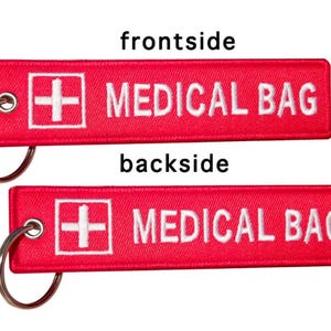 K&ouml;nnte beinhalten: Roter Stoffschl&uuml;sselanh&auml;nger mit einem wei&szlig;en Kreuz und dem Text "MEDICAL BAG" auf beiden Seiten.