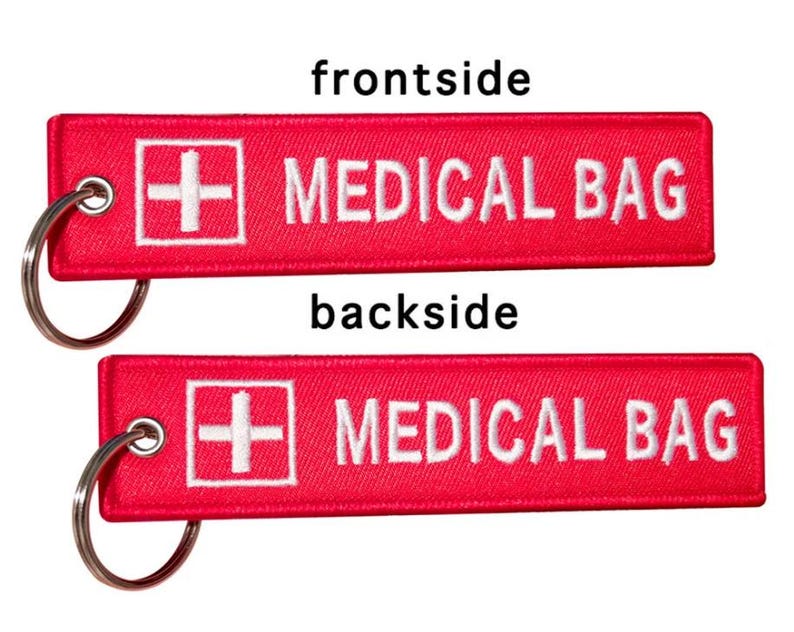 K&ouml;nnte beinhalten: Roter Stoffschl&uuml;sselanh&auml;nger mit einem wei&szlig;en Kreuz und dem Text "MEDICAL BAG" auf beiden Seiten.