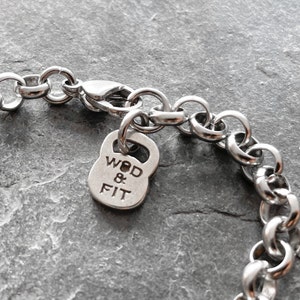 May include: A silver chain bracelet with a silver charm in the shape of a kettlebell. The charm has the words "WOD & FIT" engraved on it.