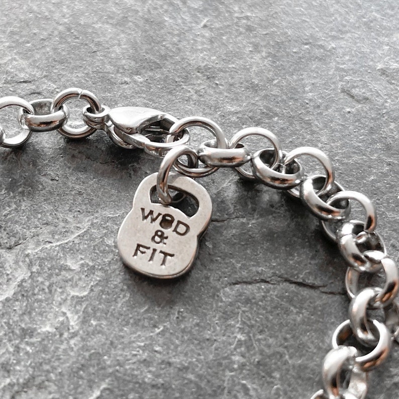 May include: A silver chain bracelet with a silver charm in the shape of a kettlebell. The charm has the words "WOD & FIT" engraved on it.