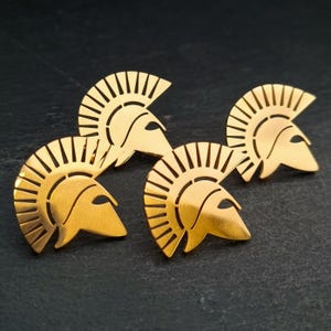 May include: Three gold-toned metal pins shaped like Spartan helmets. Each helmet features a prominent crest with vertical cutouts and a pointed visor section. The metallic finish is smooth and reflective, showcasing the distinct silhouette of the headgear.