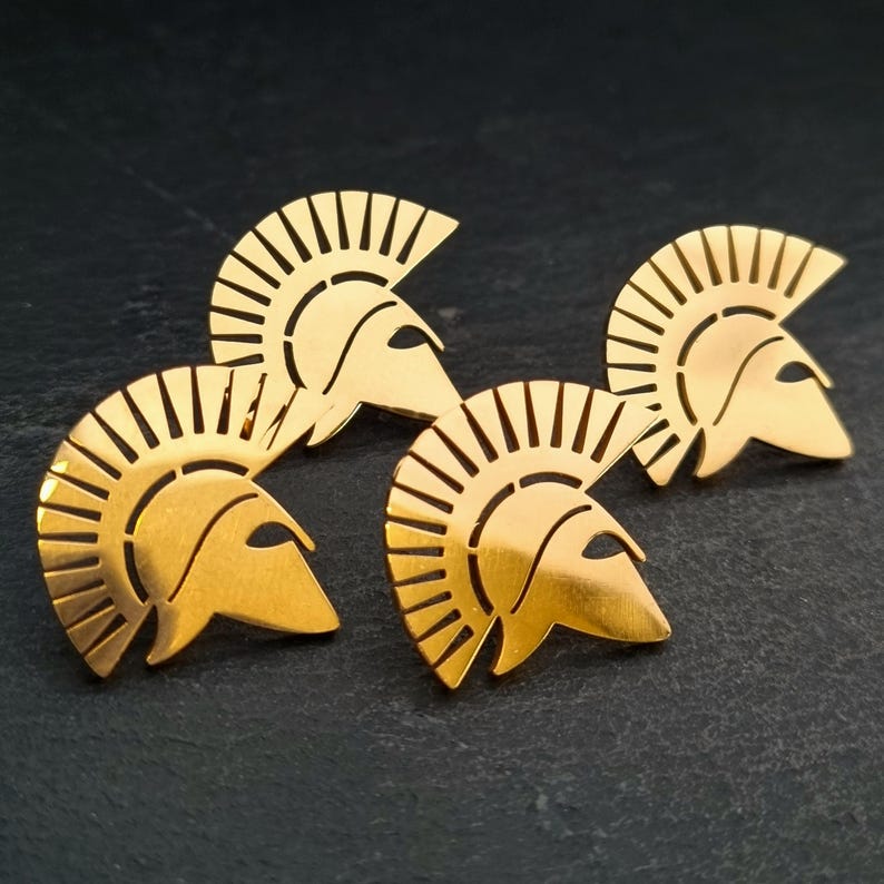 May include: Three gold-toned metal pins shaped like Spartan helmets. Each helmet features a prominent crest with vertical cutouts and a pointed visor section. The metallic finish is smooth and reflective, showcasing the distinct silhouette of the headgear.