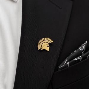 May include: A gold-toned metal lapel pin shaped like a Spartan helmet with a prominent crest. The pin is attached to the lapel of a black suit jacket, which is worn over a white shirt and accompanied by a black pocket square with a white pattern.