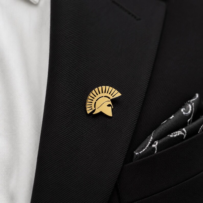 May include: A gold-toned metal lapel pin shaped like a Spartan helmet with a prominent crest. The pin is attached to the lapel of a black suit jacket, which is worn over a white shirt and accompanied by a black pocket square with a white pattern.