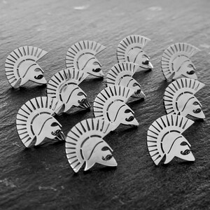 May include: A collection of ten silver-toned metal pins, each shaped like a Spartan helmet with a prominent crest and a small, distinct moustache detail on the faceplate. The pins are arranged in a scattered formation, showcasing their metallic finish.