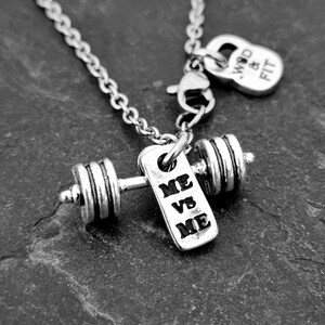 May include: Silver necklace with a dumbbell charm and a tag that reads "ME VS ME". The necklace also has a small charm that reads "WOD & FIT".