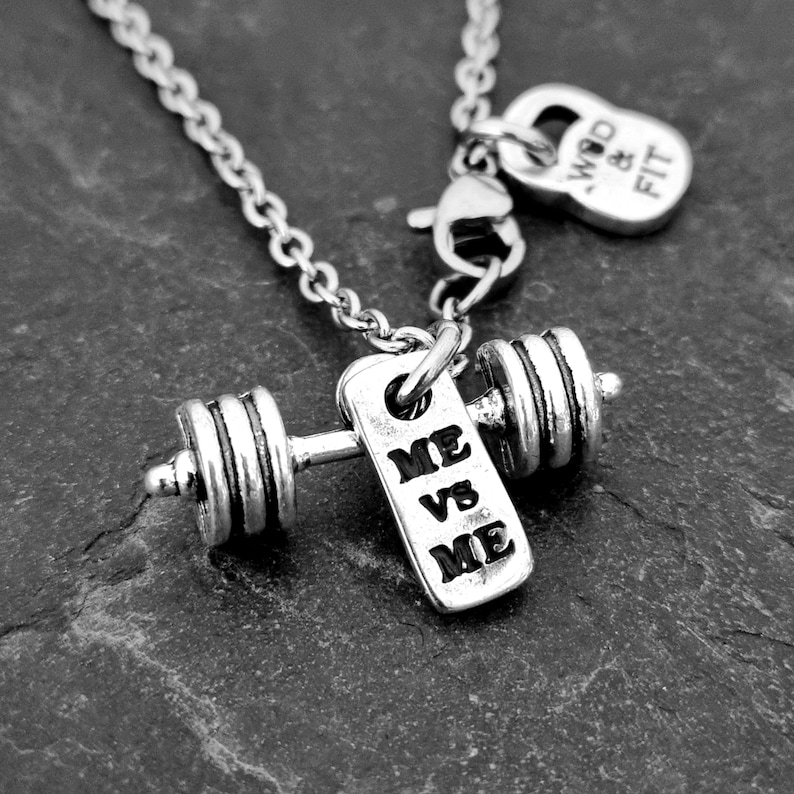 May include: Silver necklace with a dumbbell charm and a tag that reads "ME VS ME". The necklace also has a small charm that reads "WOD & FIT".
