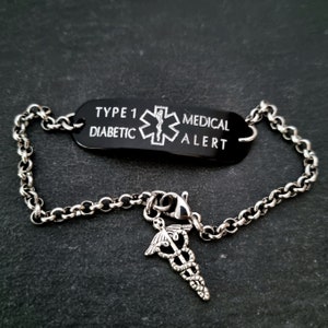 Può includere: Un braccialetto di allerta medica nero con una piastra rettangolare incisa con il testo TYPE 1 DIABETIC MEDICAL ALERT e un simbolo della Stella della Vita. Il braccialetto ha una catena color argento e un ciondolo caduceo appeso alle maglie.