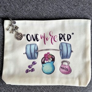 Könnte beinhalten: Cremefarbene Kosmetiktasche aus Canvas mit Fitness-Motiv. Die Tasche zeigt eine Langhantel, Kurzhanteln und Kettlebells mit dem Text "ONE MORE REP" in Schwarz und Lila. Ein Anhänger mit Miniatur-Gewichten hängt am Reißverschluss.