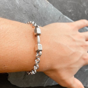 May include: A silver chain bracelet with a dumbbell charm. The dumbbell is made of two square-shaped pieces connected by a bar.