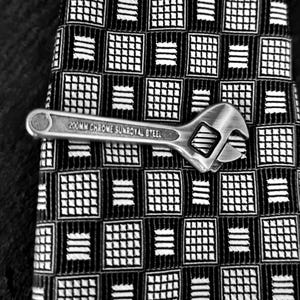 May include: A silver-coloured wrench tie clip with the text "200MM CHROME SUNROYAL STEEL" is displayed on a black and white patterned tie. The tie has a repeating square and stripe design.