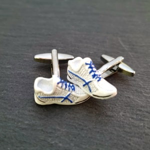 Può includere: Un paio di gemelli a forma di scarpa da corsa bianca con dettagli blu. I gemelli sono in metallo e hanno una finitura argentata.