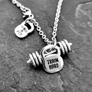 May include: Silver necklace with a kettlebell and barbell charm. The charm has the words "Train Hard" engraved on it. The necklace also has a smaller charm with the words "WOD & Fit" engraved on it.