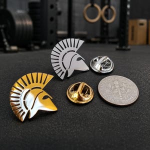 May include: Two Spartan helmet lapel pins, one in a polished silver tone and one in a polished gold tone, displayed alongside their respective metal clutch backings and a United States dime for scale on a dark, textured surface.
