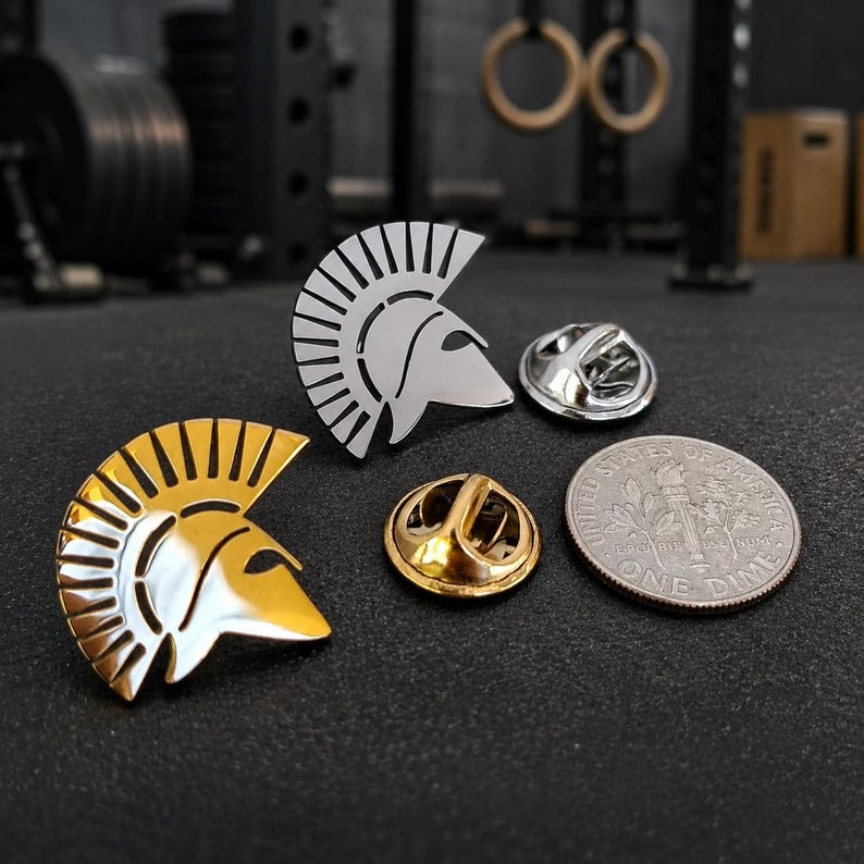 May include: Two Spartan helmet lapel pins, one in a polished silver tone and one in a polished gold tone, displayed alongside their respective metal clutch backings and a United States dime for scale on a dark, textured surface.