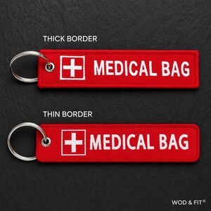 May include: Two rectangular red fabric keychains with silver metal rings. Each features a white embroidered medical cross icon and the text MEDICAL BAG in white capital letters. One keychain has a thick white border, the other a thin border.