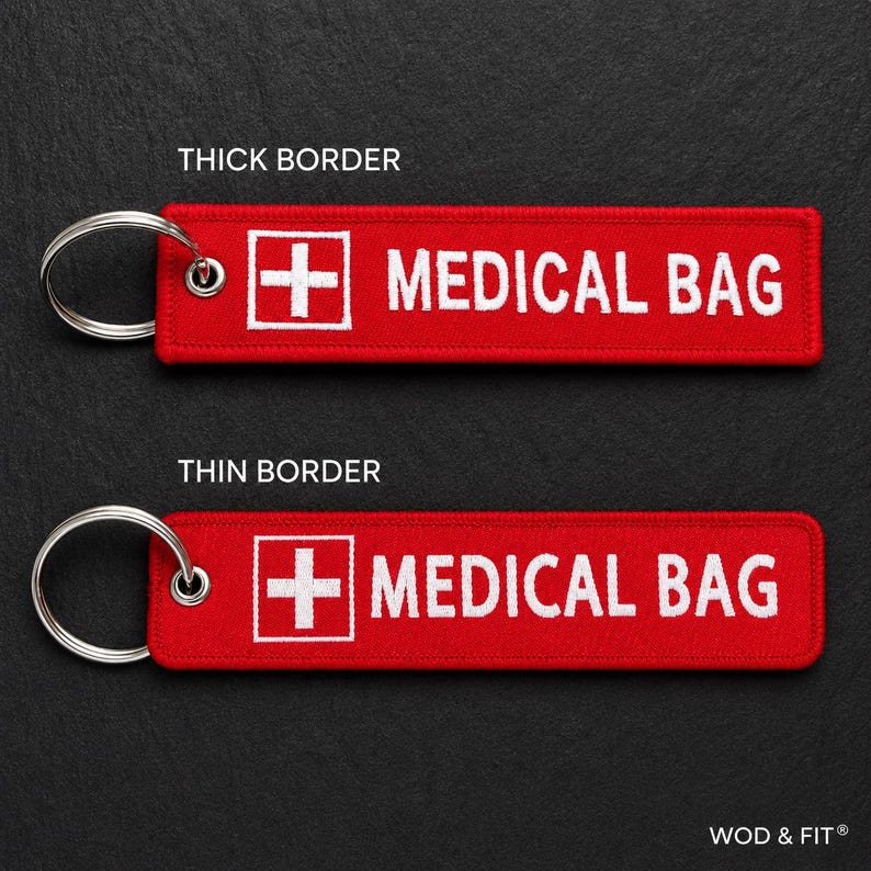 May include: Two rectangular red fabric keychains with silver metal rings. Each features a white embroidered medical cross icon and the text MEDICAL BAG in white capital letters. One keychain has a thick white border, the other a thin border.