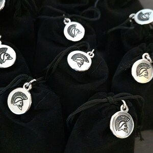 May include: A collection of small black velvet drawstring pouches, each featuring a circular silver-toned metal charm attached to the cord. The charm displays a detailed embossed profile of a helmeted warrior in a classic ancient style.