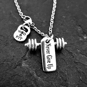 May include: Silver necklace with a dumbbell charm and a small charm that says "WOD & FIT". The dumbbell charm has a tag that says "Never Give Up".
