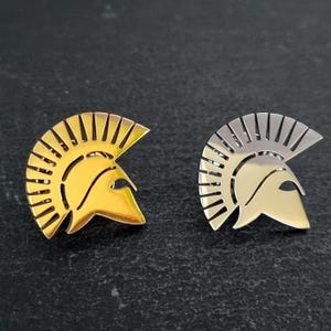 May include: Two metal lapel pins shaped like Spartan helmets. One pin has a polished gold-tone finish, and the other has a polished silver-tone finish. Both pins feature a distinct crest design with vertical cutouts along the top edge.