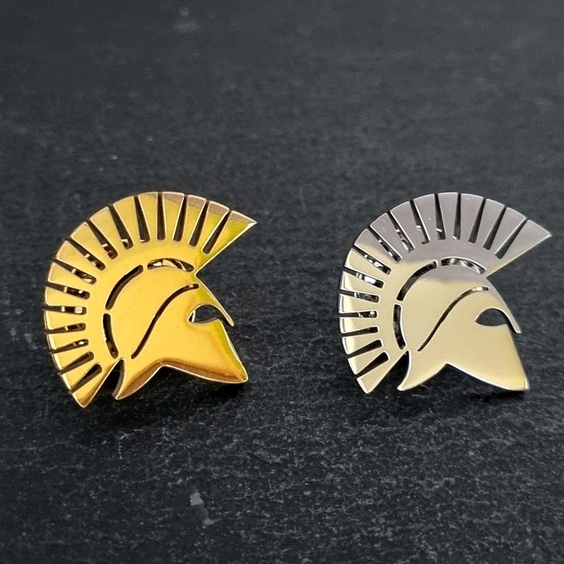May include: Two metal lapel pins shaped like Spartan helmets. One pin has a polished gold-tone finish, and the other has a polished silver-tone finish. Both pins feature a distinct crest design with vertical cutouts along the top edge.