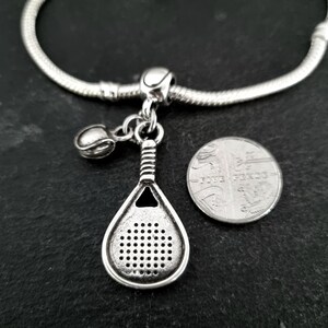 May include: Silver charm bracelet with a silver padel racket charm, a silver ball charm, and a silver bead. The charm is hanging from a silver chain.