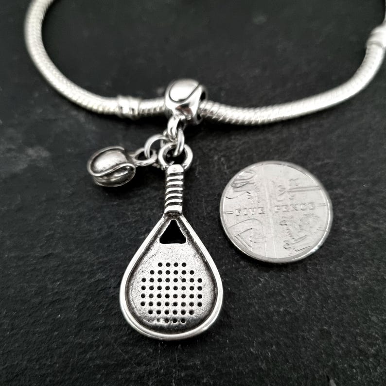 May include: Silver charm bracelet with a silver padel racket charm, a silver ball charm, and a silver bead. The charm is hanging from a silver chain.