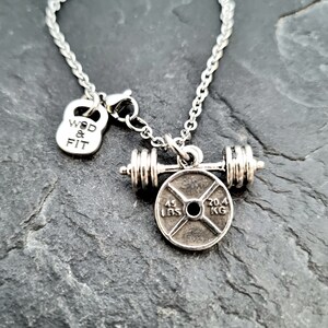 May include: Silver necklace with a weight plate charm and a barbell. The weight plate has the text "45 LBS" and "20.4 KG" on it. The charm also includes a small kettlebell with the text "WOD & FIT" on it.