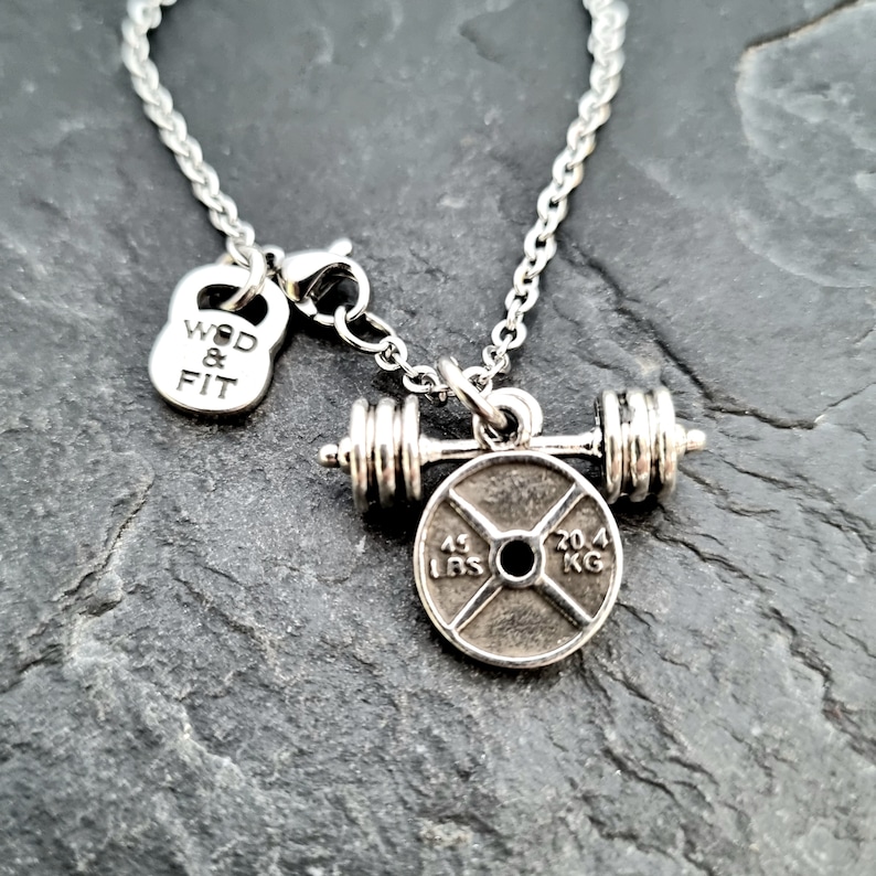 May include: Silver necklace with a weight plate charm and a barbell. The weight plate has the text "45 LBS" and "20.4 KG" on it. The charm also includes a small kettlebell with the text "WOD & FIT" on it.