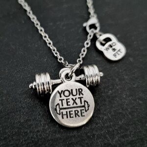May include: Silver necklace with a barbell charm and a round pendant that says "YOUR TEXT HERE". The necklace also has a small charm shaped like a kettlebell that says "WOD & FIT".